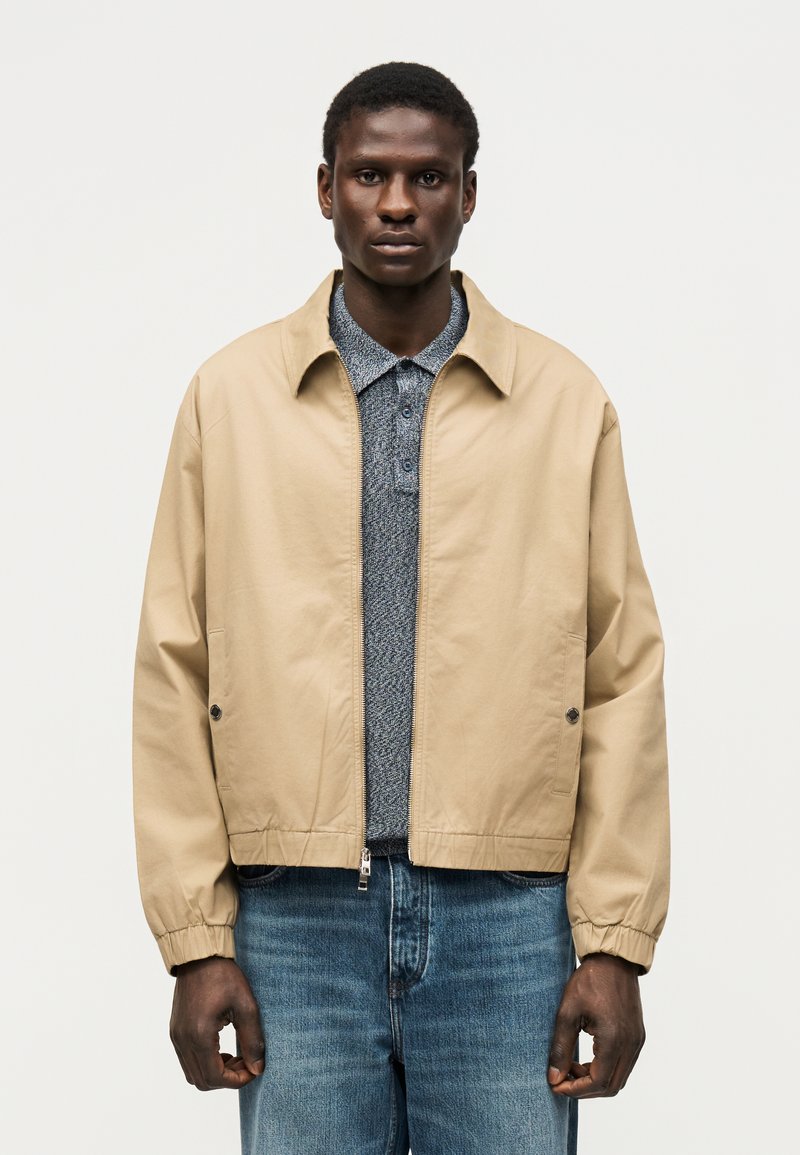 Άνδρας με μπεζ ζακέτα με φερμουάρ πάνω από μπλε πουκάμισο πόλο με σχέδια και μπλε τζιν, στέκεται μπροστά σε απλό φωτεινό φόντο.