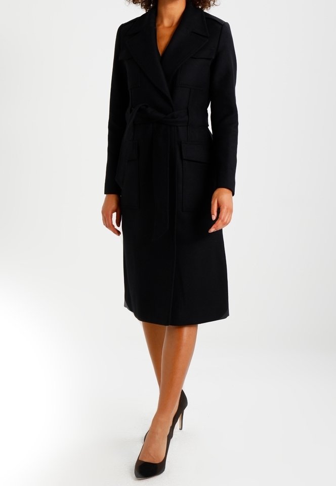 Femme portant un long manteau noir ceinturé avec des revers et des poches à rabat, assorti à des talons noirs.