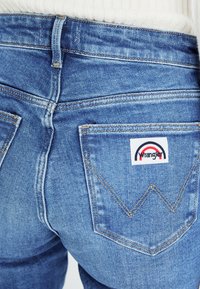 Jeans in denim blu con vita alta, tasche posteriori con etichetta "Wrangler" e dettagli di cucitura distintivi in un motivo a zigzag.