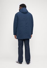 Cappotto imbottito blu navy con cappuccio, maniche lunghe e cuciture laterali. Indossato sopra pantaloni in denim scuro e sandali bianchi. Texture liscia.