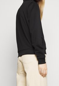 Sudadera negra ligera con cuello y puños acanalados, con bolsillos laterales y un corte relajado, combinada con pantalones beiges.