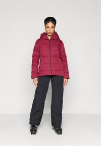 Doudoune bordeaux avec fermeture éclair à l'avant, capuche et poches latérales. Associée à un pantalon cargo noir et des bottes de ski noires. Texture lisse.