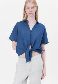Donkerblauwe spijkerhemden met korte mouwen, voorzien van een knoopsluiting aan de voorkant en een geknoopt zoom. Gecoupled met lichtgekleurde, op maat gemaakte broek.