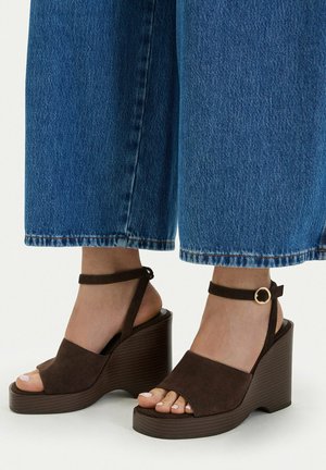 Füße, die braune Wildleder-Wedgesandalen mit Knöchelriemen und goldenen Schnallen tragen, kombiniert mit weit geschnittenen blauen Jeans.