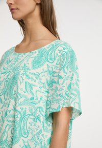 Blusa ligera de manga corta en una tela color crema con un estampado de cachemira verde, que presenta un escote redondeado y un corte holgado.