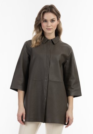 Giovane donna con capelli mossi fino alle spalle che indossa un cappotto di pelle marrone scuro abbottonato con maniche tre quarti e pantaloni chiari.