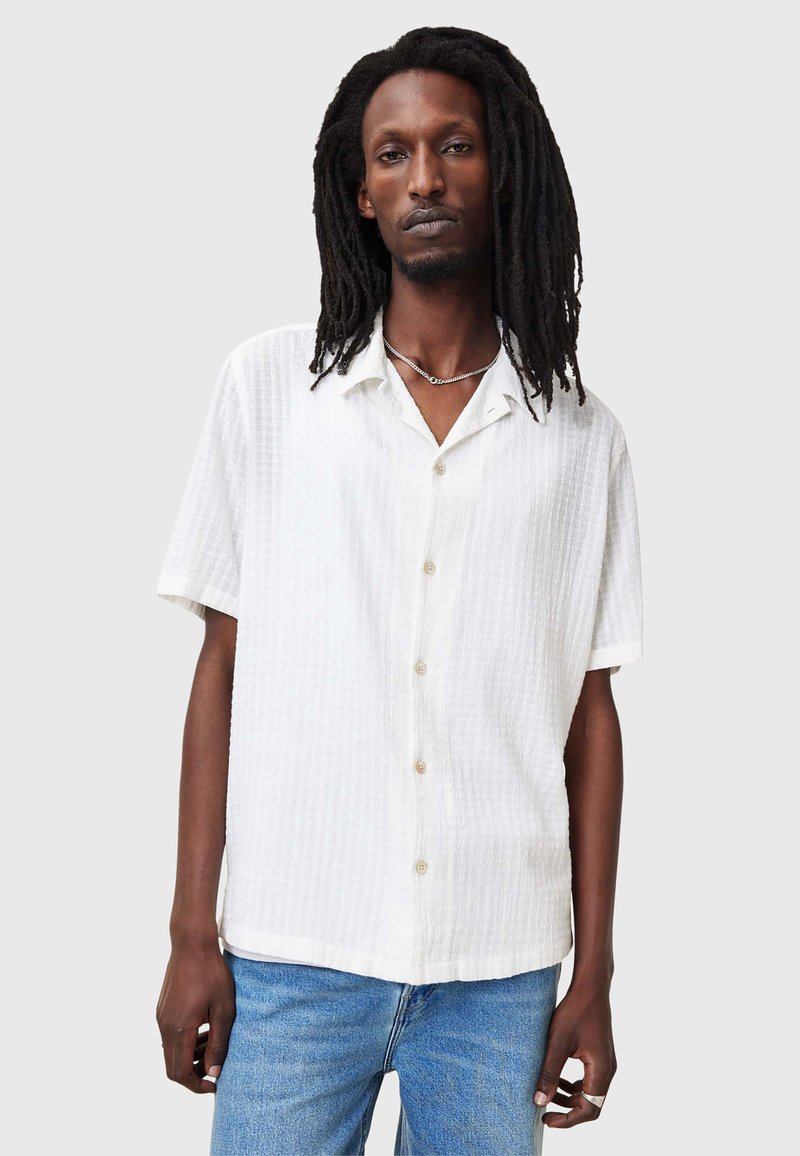 Camicia bianca a maniche corte in tessuto strutturato, con bottone frontale e colletto. Abbinata a jeans in denim blu.