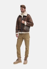 Chaqueta de shearling de cuero marrón con cierre de cremallera, cuello crema, pantalones cargo beige y botas marrones, llevando un bolso texturizado marrón.