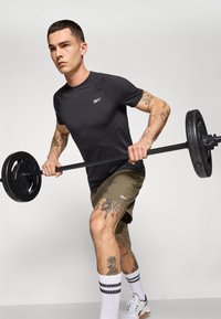 T-shirt de sport à manches courtes noir, shorts vert olive, tenant une haltère. Tatouages visibles sur les bras et les jambes, chaussettes blanches avec des rayures noires, chaussures de sport.