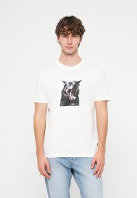 Balts kokvilnas T-krekls ar melna suņa, kas rēc, grafiku. Apaļa kakla izgriezums un īsas piedurknes. Smalks zīmola logo attēla apakšdaļā.