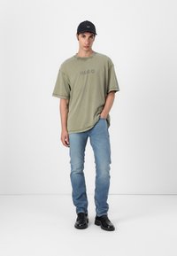 Olive green oversized t-shirt with "HUGO" logo, light blue jeans, and black cap. Black leather shoes complete the outfit.