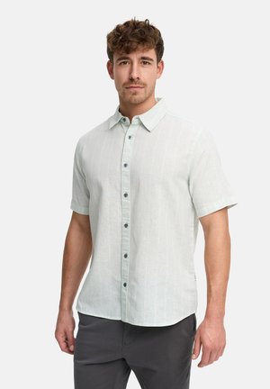 Mand med krøllet hår iført en lys kortærmet skjorte med knapper og mørke bukser, stående foran en ensfarvet baggrund.