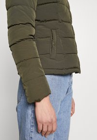 Oliven grøn puffjakke med horisontalt quiltet mønster, med sidelomme med knap og ribstrikkede manchetter, kombineret med lyseblå jeans.