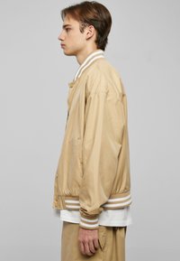 Giubbotto bomber leggero beige con colletto e polsini a strisce bianche e marroni, dotato di bottoni a pressione e tasche laterali. Texture liscia.