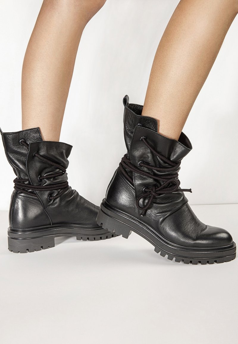 Bottes cheville en cuir noir avec semelles épaisses et lacets enroulés autour du haut, portées sur des jambes nues sur un fond uni.