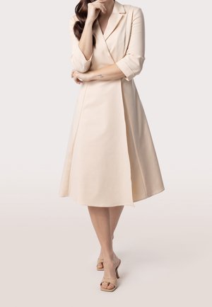 Kvinde iført en beige, knælængde, langærmet slå-om-kjole med revers, stående med den ene arm over kors og hånden tæt på ansigtet, beige høje hæle.