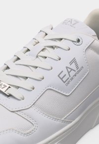 Λευκό αθλητικό παπούτσι EA7 Emporio Armani με λευκούς κορδόνια, πάνελ από δίχτυ και δέρμα, και μεταλλική ταμπέλα με το λογότυπο μπροστά.