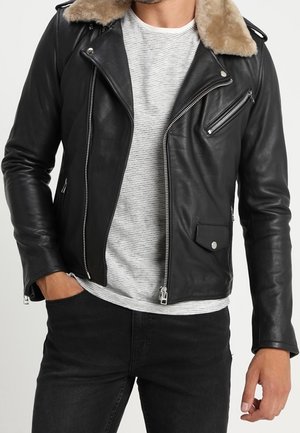 Mann trägt eine schwarze Leder-Bikerjacke mit beigem Fellkragen über einem weiß-grau gestreiften Hemd, kombiniert mit schwarzen Jeans.