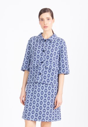 Donna che indossa una giacca con bottoni a motivi blu e bianchi con maniche a gomito e una gonna abbinata al ginocchio, in piedi su sfondo bianco.