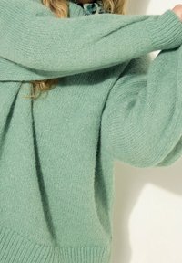 Donna che indossa un morbido maglione lavorato a maglia verde menta con maniche lunghe e leggermente a sbuffo, mentre posa con le braccia incrociate vicino al viso.