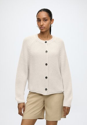 Donna che indossa un cardigan in maglia color crema con bottoni e pantaloncini beige, in piedi di fronte con espressione neutra su uno sfondo semplice.