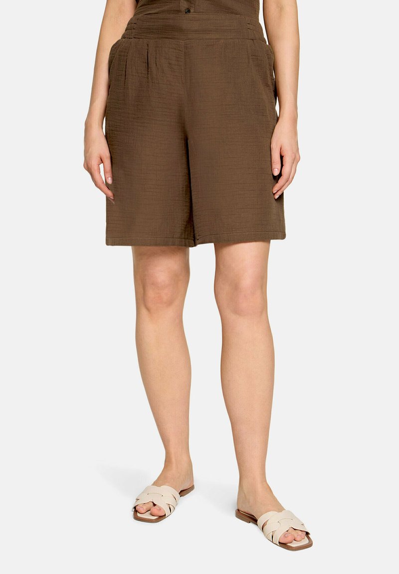 Personne portant un short marron mi-long avec des poches latérales et des sandales plates beiges, debout devant un fond uni.