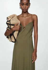 Olivově zelené šaty s přiléhavým živůtkem, detailem ve tvaru klíčové dírky a nastavitelnými ramínky. Modelka drží béžovou háčkovanou kabelku s černými akcenty.