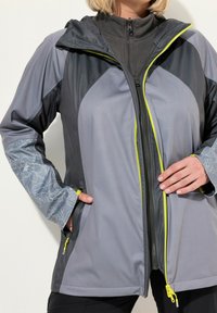 Veste multi-panneaux grise avec capuche, fermeture éclair à l'avant, manches à motif floral et zips jaunes. Le matériau semble lisse et léger.
