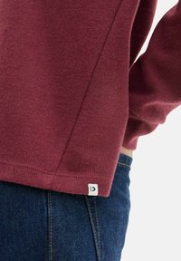 Bordo džemperis, izgatavots no mīksta adīta auduma, ar brīvu piegriezumu, smalku tekstūru un baltu etiķeti ar burtu apakšmalā.