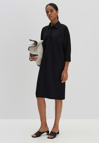 Schwarzes Hemdkleid mit Kragen und Dreiviertelärmeln, kombiniert mit flachen schwarzen Mules und einer weißen und grauen strukturierten Tasche.