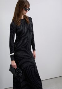 Abito lungo nero con motivi grigi vorticosi, realizzato in tessuto strutturato. Presenta maniche lunghe e un braccialetto d'argento. Tiene in mano una pochette nera.