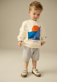 Sweatshirt crème avec un graphique coloré, shorts rayés et chaussures grises avec des sangles Velcro, mettant en avant un style décontracté pour enfants.