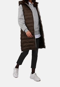 Person wearing a long brown padded vest over a gray hoodie, black cropped pants, black tights, and white sneakers walking forward.