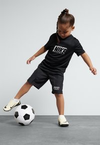 Black Nike t-shirt and shorts. Child is balancing a black and white soccer ball with one foot, wearing cream-colored soccer shoes.