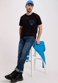 Czarna koszulka z niebieskim logo, niebieska czapka, niebieski sweter zarzucony na białe krzesło, dżinsy oraz czarne buty z teksturowaną podeszwą.