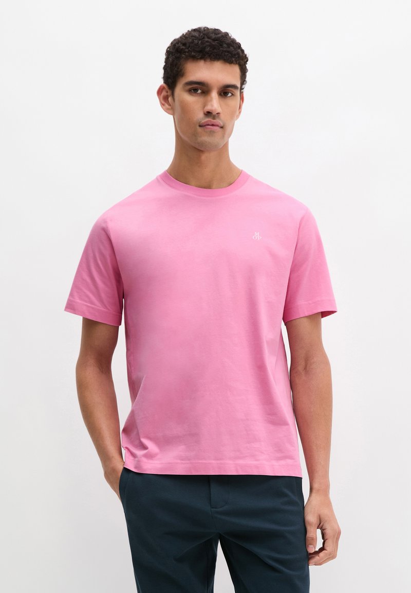 Νεαρός άνδρας με κοντά σγουρά μαλλιά, φορώντας απλό ροζ κοντομάνικο μπλουζάκι και σκούρο παντελόνι, στέκεται με το ένα χέρι στην τσέπη.