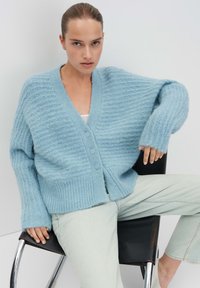 Cardigan lavorato a maglia azzurro chiaro con scollo a V, tre bottoni e polsini a coste. Vestibilità ampia e strutturata, indossato sopra una maglietta bianca e jeans chiari.