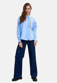 Femeie purtând o cămașă largă albastru deschis cu nasturi și detaliu text, combinată cu blugi evazați de culoare albastru închis, stând cu o mână relaxată.