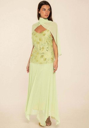 Mujer con camiseta sin mangas de flores verde pálido con falda fluida a juego y chal ligero sobre los hombros, de pie frente a un fondo liso.