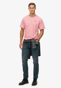 Roze katoenen t-shirt met korte mouwen, gecombineerd met donkerblauwe jeans en een zwart-wit geruit shirt om de taille geknoopt. Zwarte schoenen.