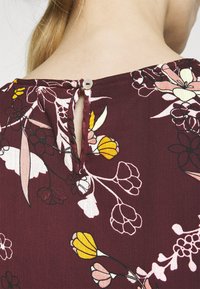 Viininpunainen kukkakuosinen pusero, jossa on nappikiinnitys takana. Siinä on avainreikädetaili ja sileä, teksturoitu kangas, jossa on erilaisia kukkakuvioita.