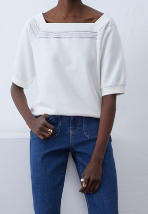 Personne portant un chemisier blanc avec des détails en broderie près de l'encolure carrée et un jean en denim bleu, avec des bagues aux deux mains.