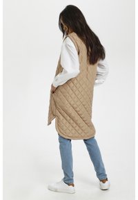 Beige quilted sleeveless vest with a high neckline, loose fit, side pockets, and a rounded hem; worn over a white long-sleeve shirt and blue jeans.