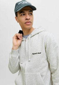 Hellgraue Kapuzenjacke mit einem weichen Textil, ausgestattet mit einem schwarzen gestickten Logo. Eine grüne Kappe mit weißem Text ergänzt das Outfit.