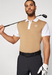 Vīriešu golfa polo krekls, brūnas krāsas ķermenis ar baltām piedurknēm, īss apkakle, priekšējais logo. Melna josta, apvienota ar tumšām biksēm un baltu golfa cimdiem.