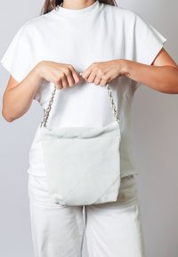 Borsa a tracolla di colore grigio chiaro, trapuntata, con una tracolla a catena tenuta in mano, caratterizzata da una texture morbida e un design angolare.