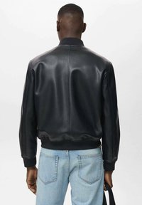 Chaqueta bomber de cuero negro con cuello y puños acanalados, que presenta una textura suave y una silueta ajustada. Llevada con unos jeans azul claro.