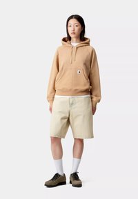 Sudadera con capucha beige y un bolsillo canguro, combinada con pantalones cortos de mezclilla claros y zapatos oscuros. Calcetines blancos completan el atuendo.