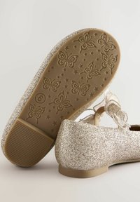 Glittriga guld ballerina skor med platt sula, med en texturerad gummisula med fjäril- och stjärnmönster, samt ett dekorativt band.