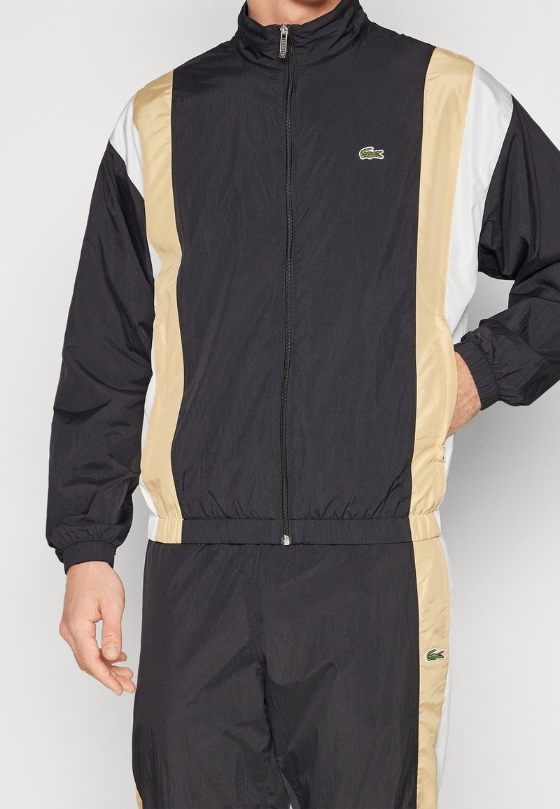 Homme portant un survêtement zippé noir, beige et blanc avec des poignets élastiques et des logos Lacoste sur la poitrine de la veste et la cuisse du pantalon.